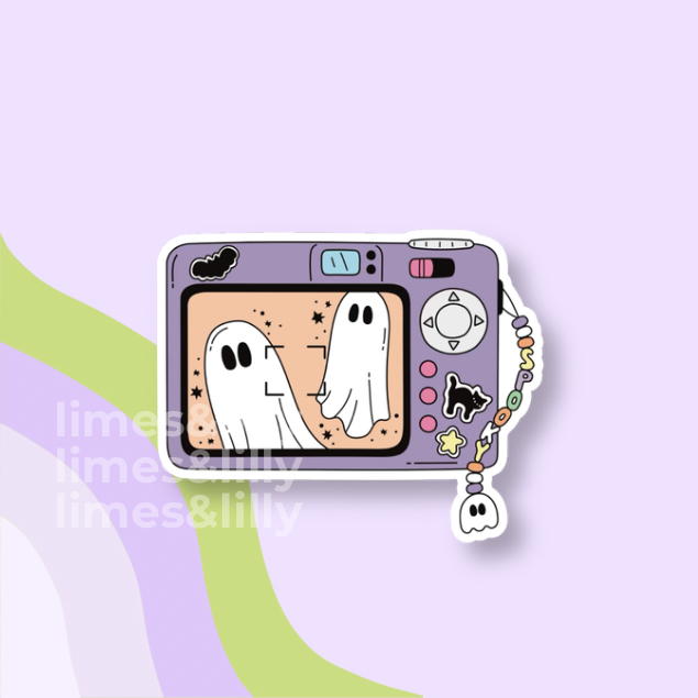 A die-cut sticker shaped like a retro digital camera, featuring a lavender body and a screen showing two white ghosts. A beaded wrist strap with a ghost charm hangs from the side.