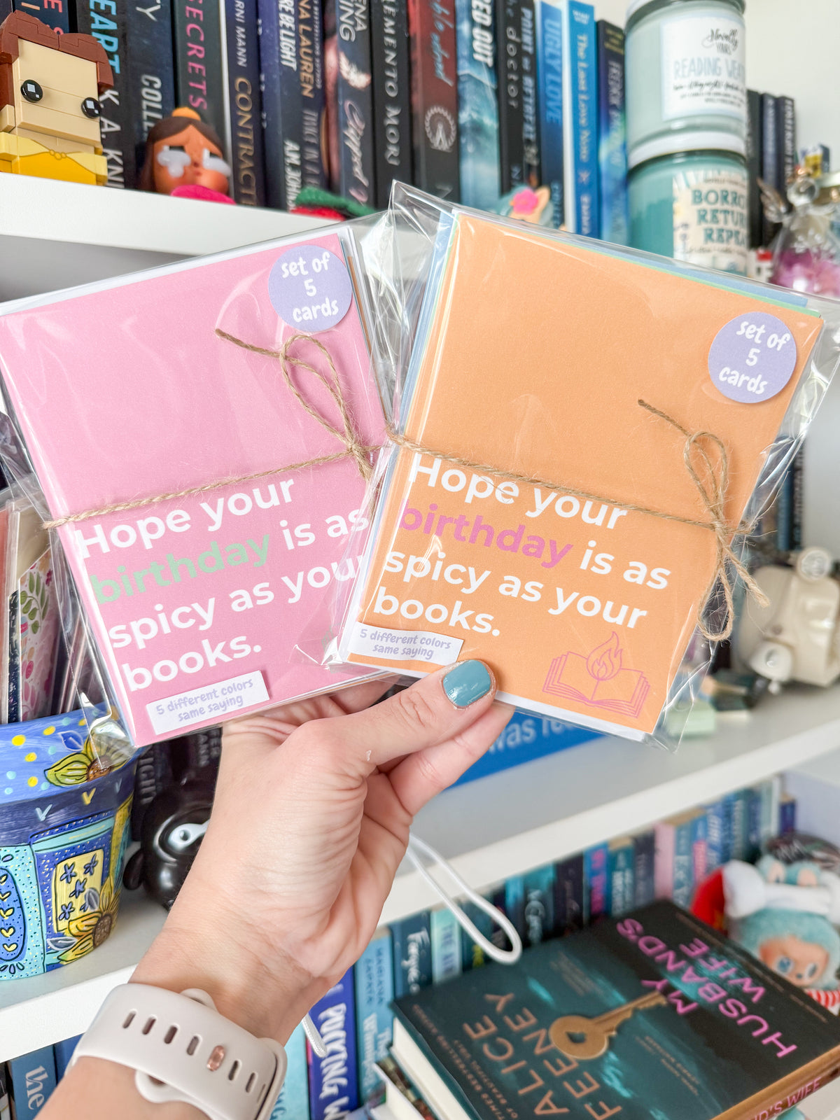 A hand holds two small packaged card sets in front of a bookshelf. One card set is pink and the other is peach/orange, both with white text reading 'Hope your birthday is as spicy as your books.