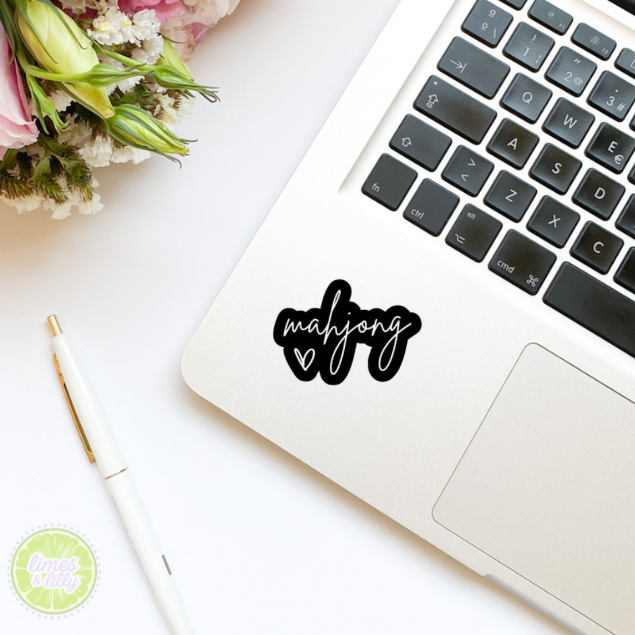 Laptop with 'mahjong' sticker on a white surface with flowers and a pen.