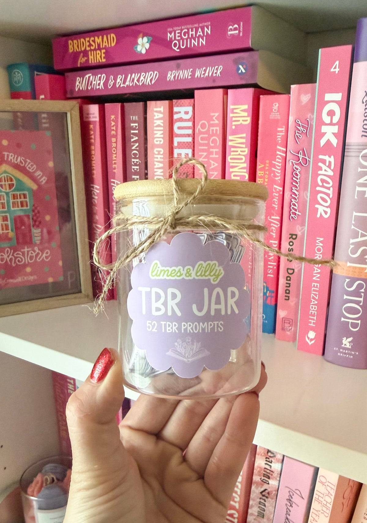 Hand holding a compact glass TBR jar filled with reading prompts in front of a bookshelf