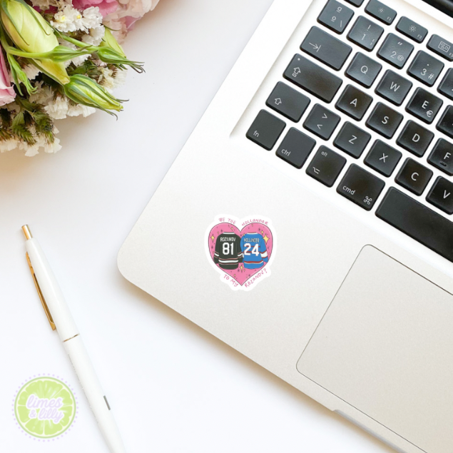 Laptop with a heart-shaped sticker on a white surface with flowers and a pen.