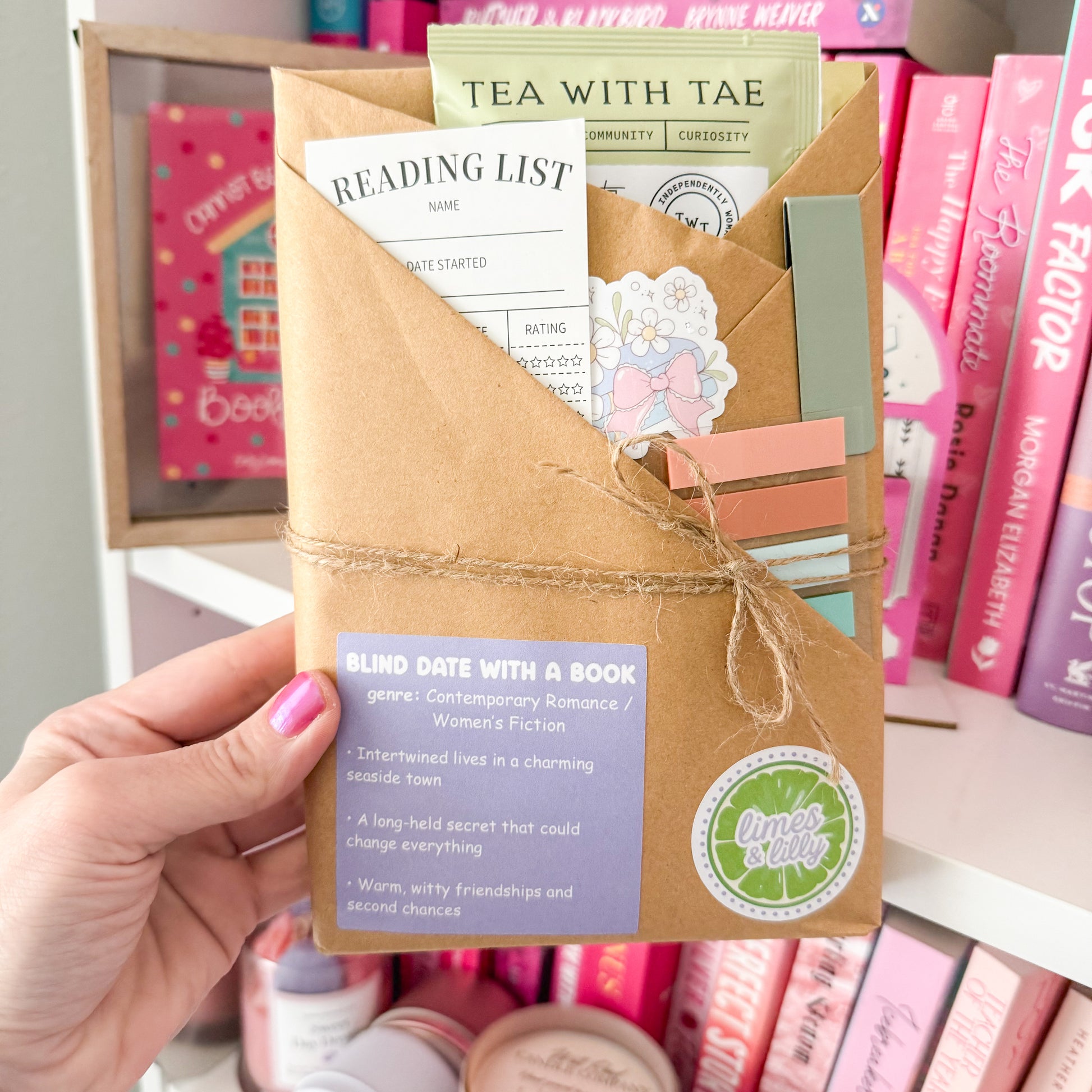 Hand holding a 'Blind Date with a Book' kit with stationery items against a bookshelf background.