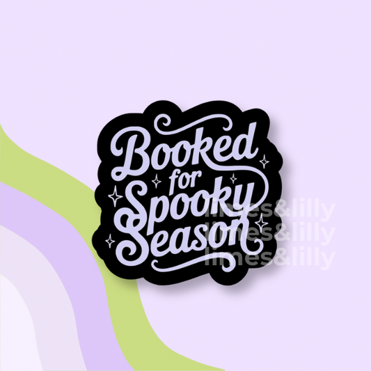 A die-cut sticker with the phrase 'Booked for Spooky Season' in white script on a black background, shown on a pastel lavender backdrop with abstract green and purple curved shapes.