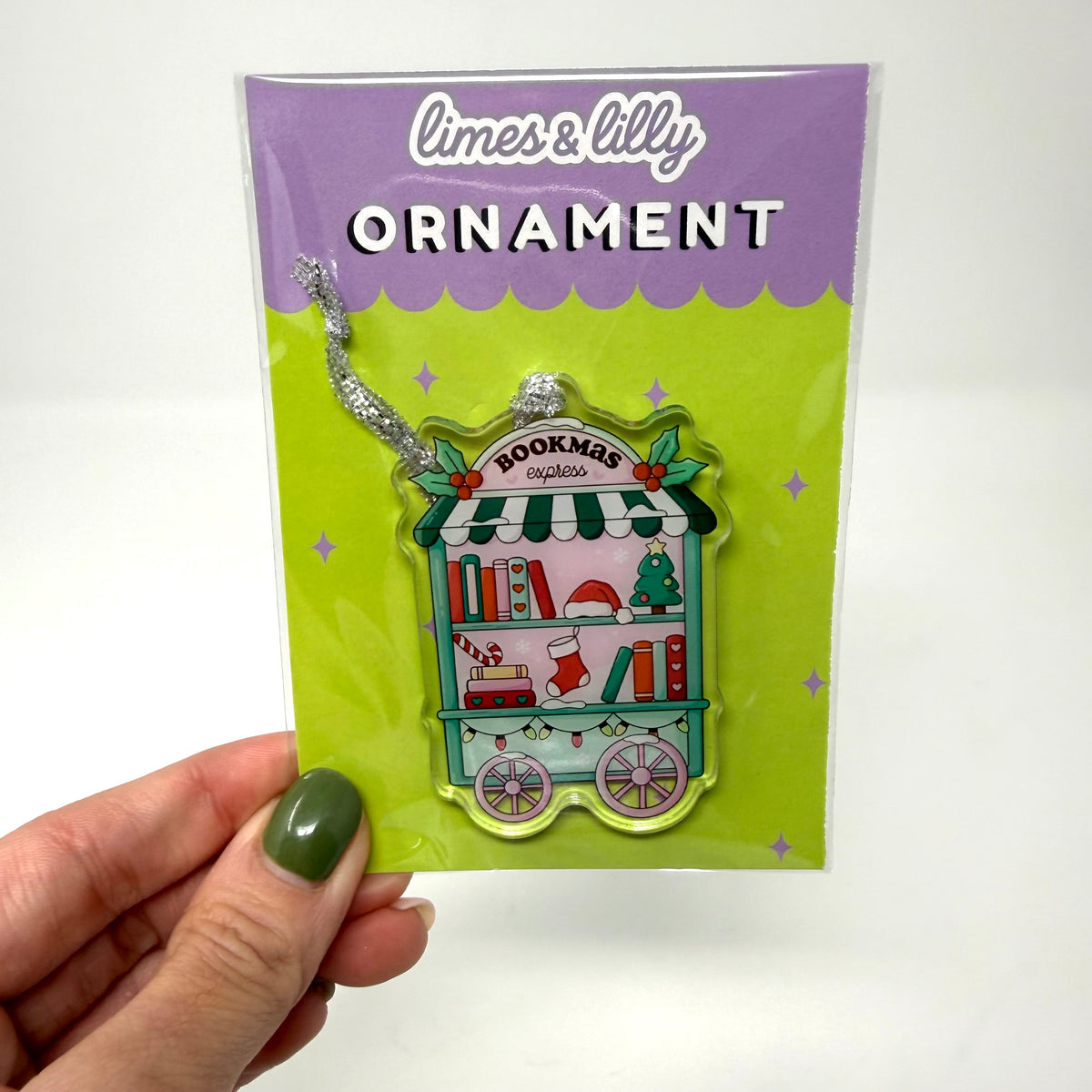 A hand with green nail polish holds a packaged acrylic Christmas bookmobile ornament. The product is in its original packaging with a purple and lime green card labeled 'limes & lilly ORNAMENT'.