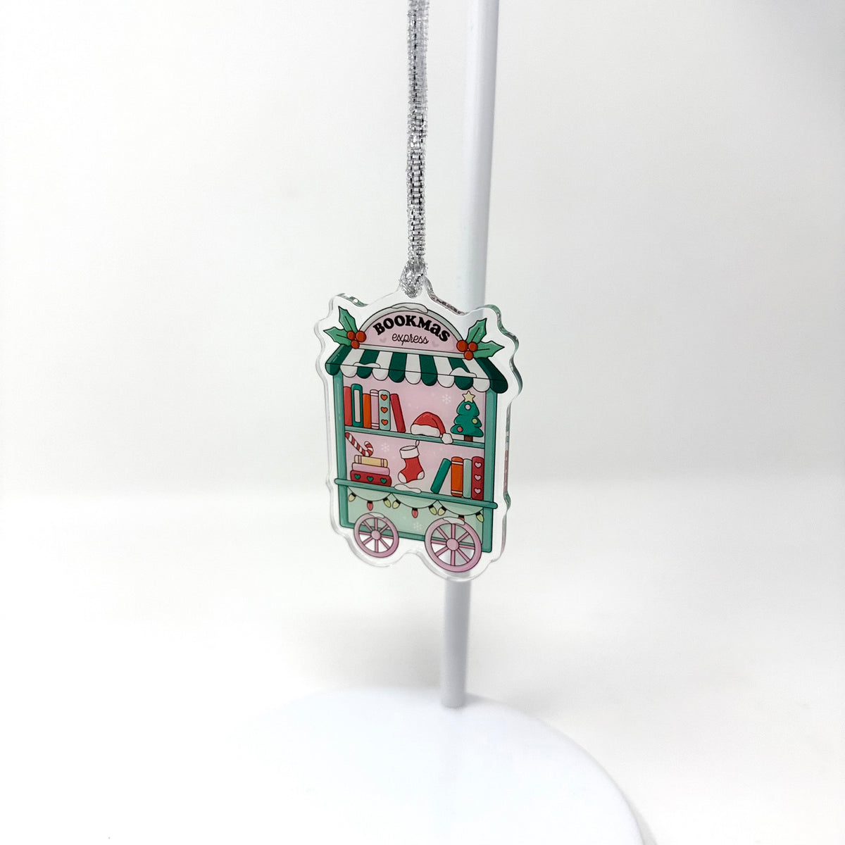 A small illustrated hanging ornament shaped like a bookish cart or stall, suspended from a silver chain on a white display stand against a white background. The ornament has a multicolor holiday design with green, red, pink, and white details and the word 'BOOKMALL' visible at the top.