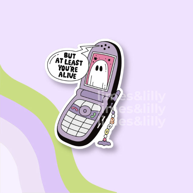 A die-cut sticker featuring a retro flip phone with a ghost on the screen and a speech bubble that reads 'BUT AT LEAST YOU'RE ALIVE'. The sticker is shown on a pastel purple background with wavy green and lavender stripes and a faint 'limes&lilly' watermark.