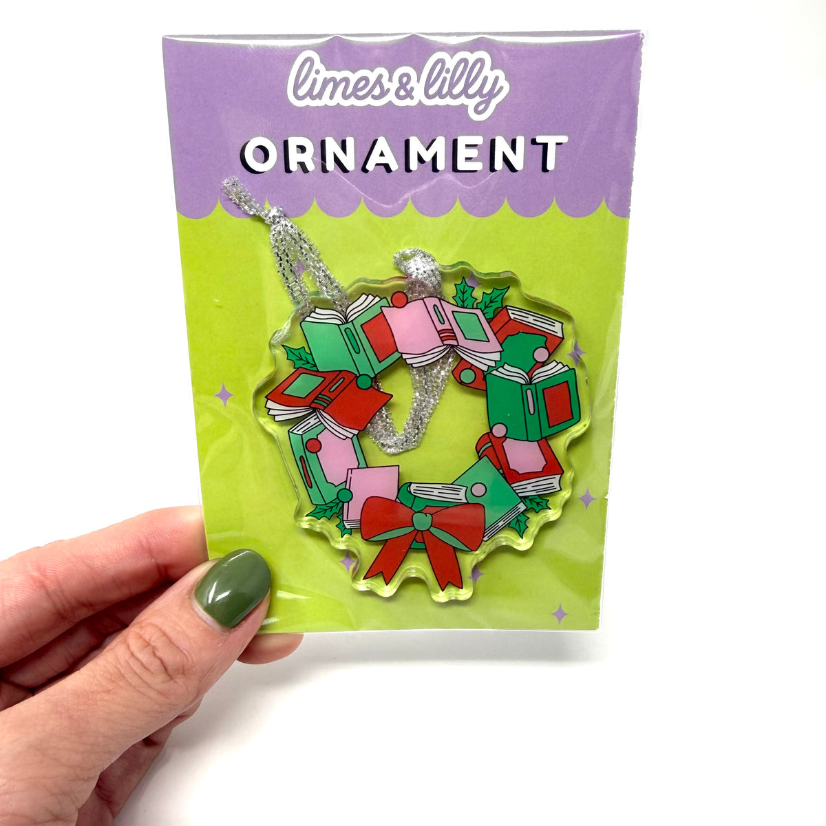 A hand with olive green nail polish holds a packaged Christmas ornament. The product is a limes & lilly ornament shaped like a wreath made of stacked presents, decorated with red, green, and pink details and a silver hanging loop, presented in its original packaging.