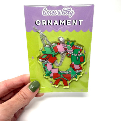 A hand with olive green nail polish holds a packaged Christmas ornament. The product is a limes & lilly ornament shaped like a wreath made of stacked presents, decorated with red, green, and pink details and a silver hanging loop, presented in its original packaging.