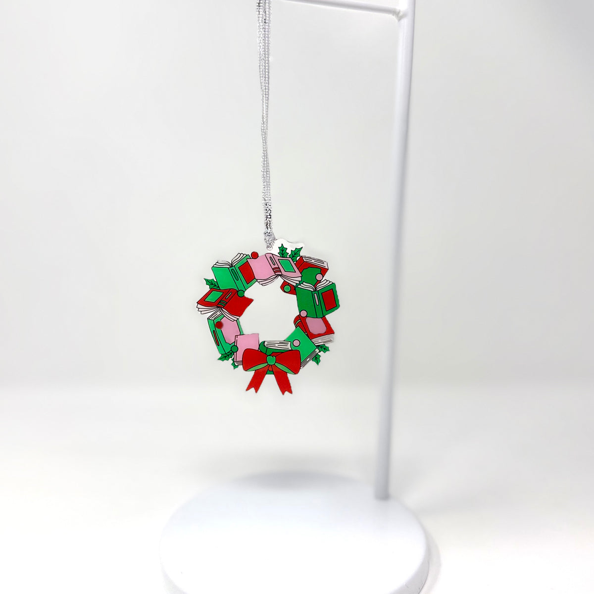 A small Christmas wreath ornament made from stacked gift boxes in red, green, and pink, hanging from a silver string on a white display stand against a white background.