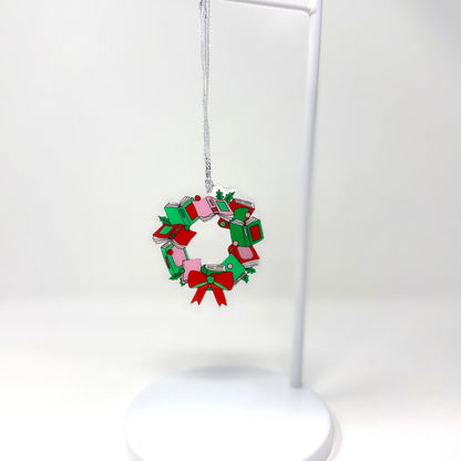 A small Christmas wreath ornament made from stacked gift boxes in red, green, and pink, hanging from a silver string on a white display stand against a white background.