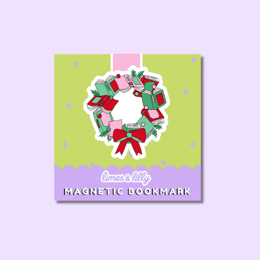 A square product card for a magnetic bookmark featuring a cute illustration of a wreath made of stacked gift boxes with a red bow. The card has a light green center, a lavender bottom band with the text 'limes & lilys MAGNETIC BOOKMARK', and is set against a solid lavender background.