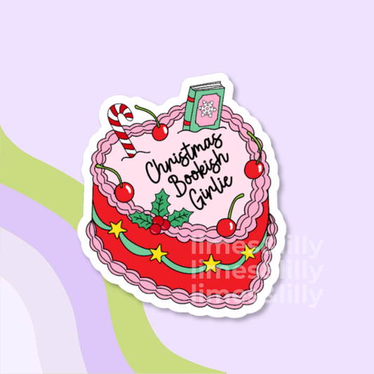A die-cut sticker featuring a whimsical illustration of a red cake with pink frosting, cherries, holly, a candy cane, a green book, and the text 'Christmas Boobeshow Girlie' in black script, shown on a pastel lavender background with abstract green and purple shapes.