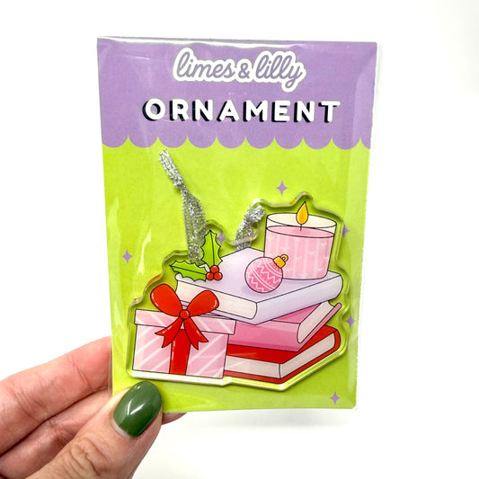 A hand with green nail polish holds a packaged Christmas ornament by limes & lily. The ornament features an illustration of stacked books, a wrapped gift with a red bow, a pink candle, a pink ornament, holly, and silver glittery accents, all on a lime green and purple backing card.