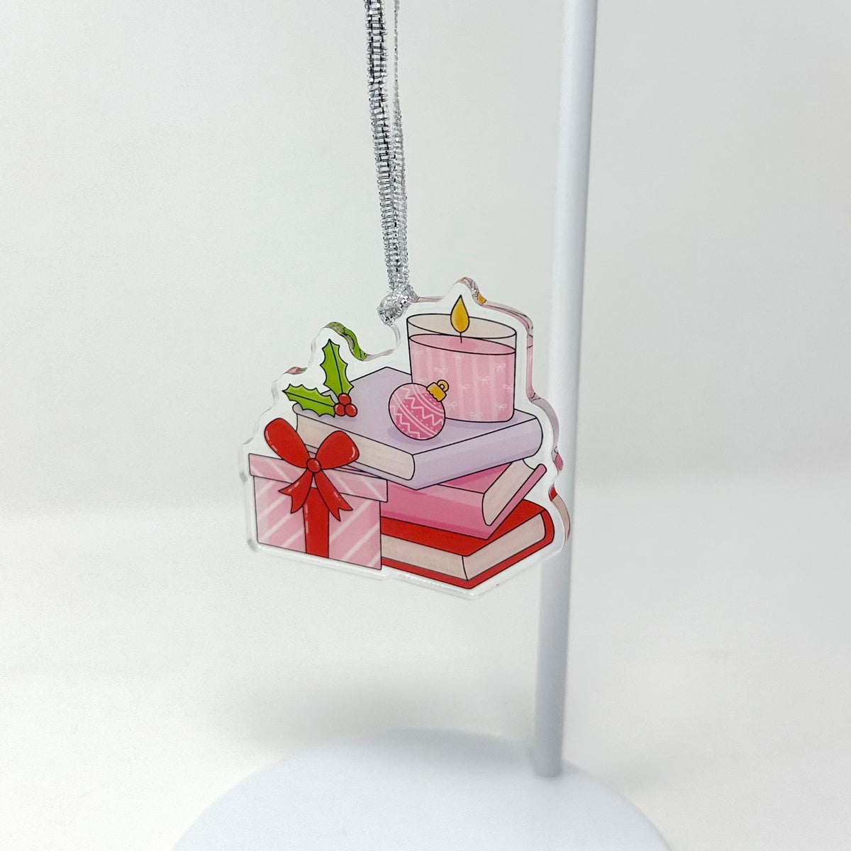 A clear acrylic Christmas ornament featuring a stack of pink books, a wrapped gift box with a red bow, a pink candle, holly, and a pink ornament, hanging from a silver string on a white display stand.