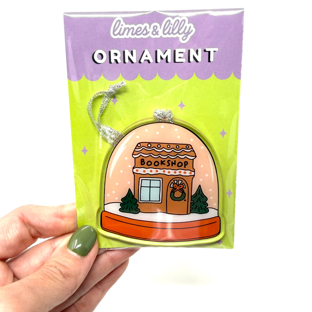 A hand with olive green nail polish holds a packaged Christmas ornament. The product is a limes & lilly brand bookshop ornament, shown in its original packaging with a bright lime green and purple card backing.