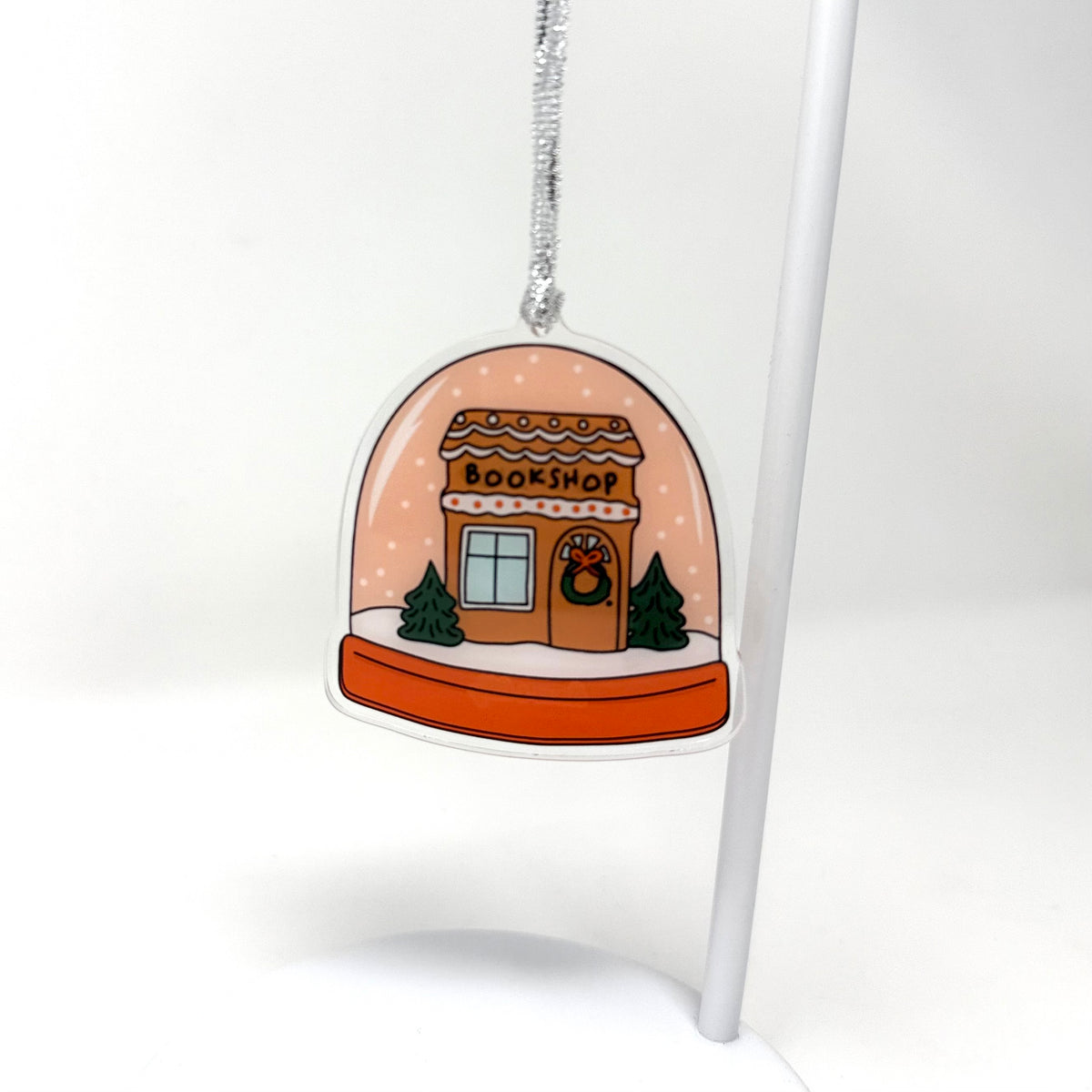 A die-cut acrylic Christmas ornament shaped like a cozy bookshop under a snowy dome, hanging from a silver string on a white display stand against a white background.