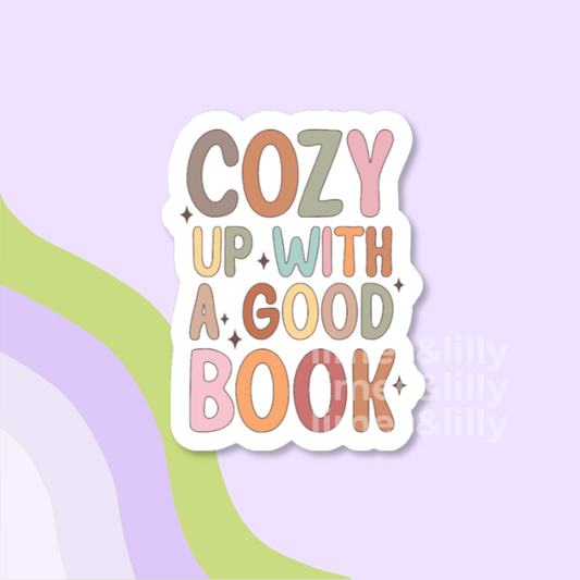 A die-cut sticker with the phrase 'COZY UP WITH A GOOD BOOK' in retro multicolor lettering on a white background, shown on a pastel lavender backdrop with abstract green and lavender curved shapes.