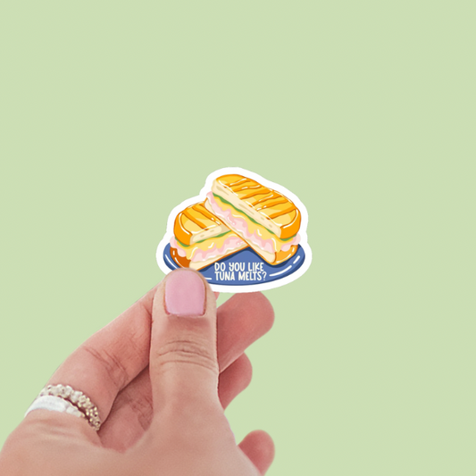 Hand holding a sticker of a tuna melt with text 'Do you like tuna melts?' against a light green background