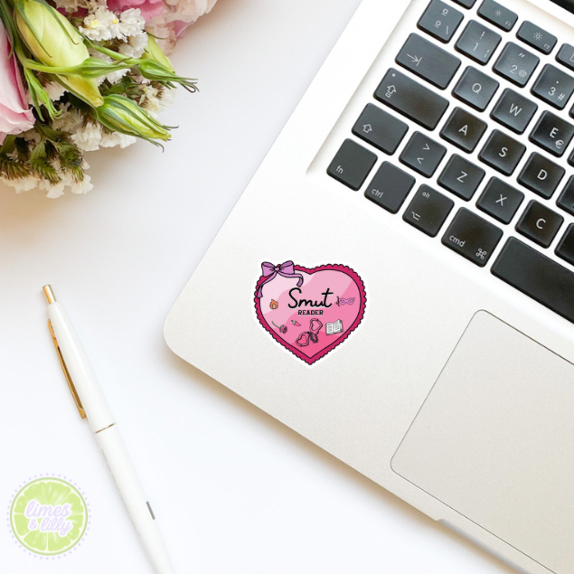 Laptop with a pink heart-shaped sticker on a white surface with flowers and a pen.