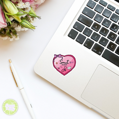 Laptop with a pink heart-shaped sticker on a white surface with flowers and a pen.