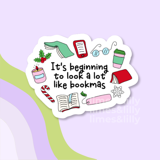 A die-cut sticker with a white border featuring cozy book-themed illustrations and the text 'It's beginning to look a lot like bookmas' in black handwritten-style font, shown on a pastel lavender background with abstract curved stripes.