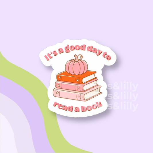 A die-cut sticker featuring the phrase 'It's a good day to read a book!' in pink lettering around a stack of three books topped with a small orange pumpkin, shown on a pastel lavender background with abstract green and lavender curved shapes.