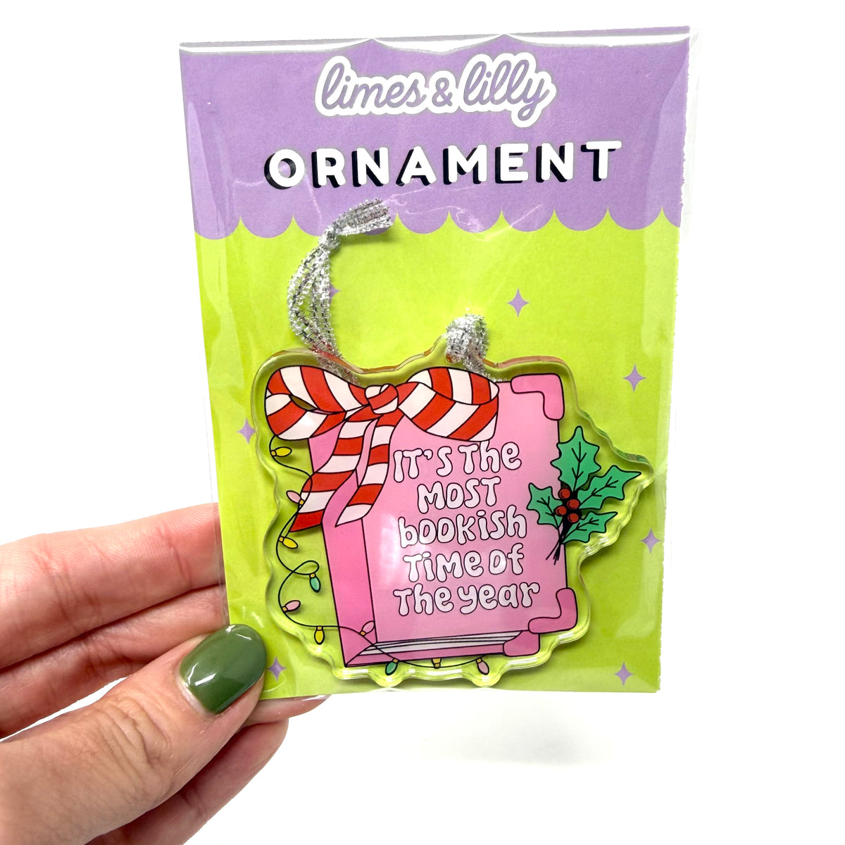 A hand with green nail polish holds a limes & lilly Christmas ornament in its original packaging. The ornament is shaped like a pink book with a red and white striped bow, holly, and the text 'It's the most bookish time of the year'.