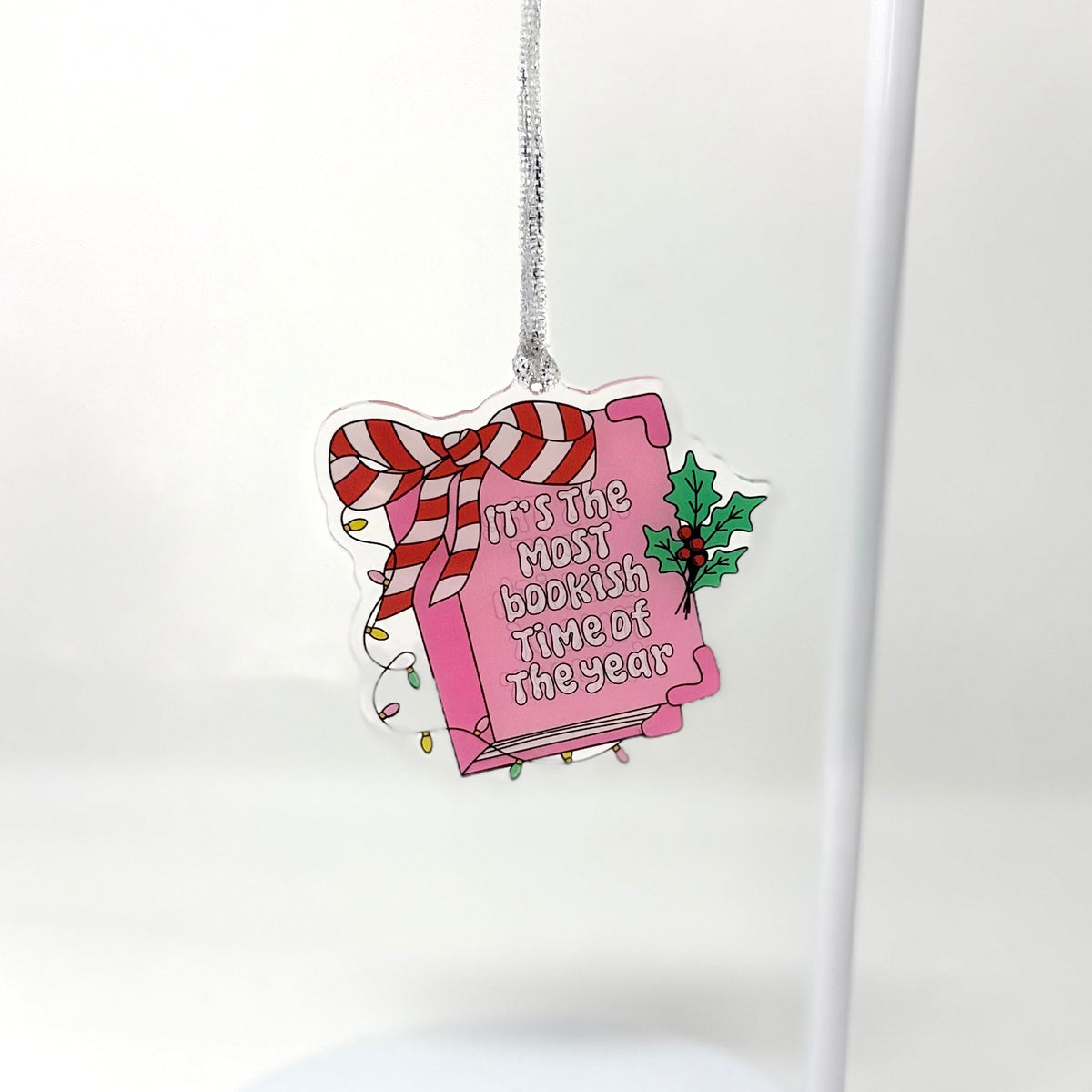 A die-cut acrylic Christmas ornament shaped like a pink book tied with a red-and-white striped bow, hanging from a silver string against a clean white background.