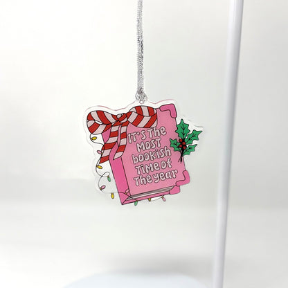 A die-cut acrylic Christmas ornament shaped like a pink book tied with a red-and-white striped bow, hanging from a silver string against a clean white background.
