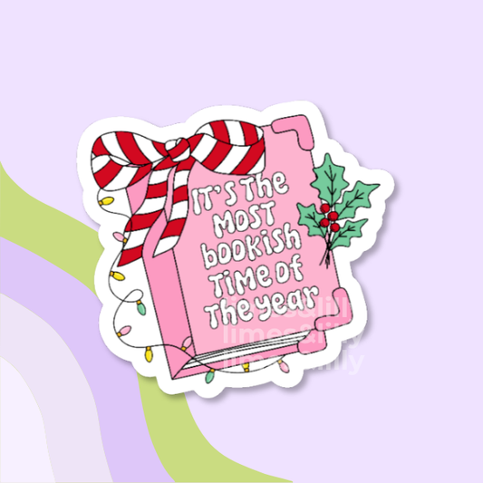 A die-cut sticker featuring a pink book wrapped in a red-and-white striped ribbon, with the phrase “It’s the most bookish time of the year” in white lettering, plus green holly leaves and colorful string lights, shown on a pastel lavender background.