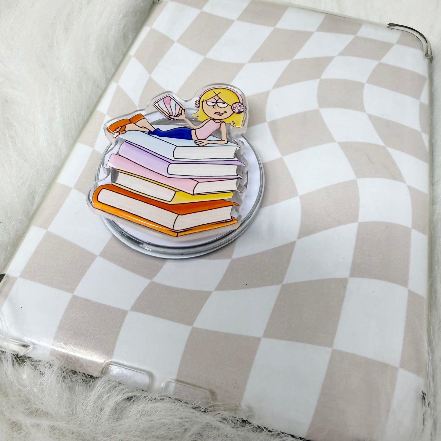 Pin with a design of a person reading a book on a stack of books, placed on a checkered surface.
