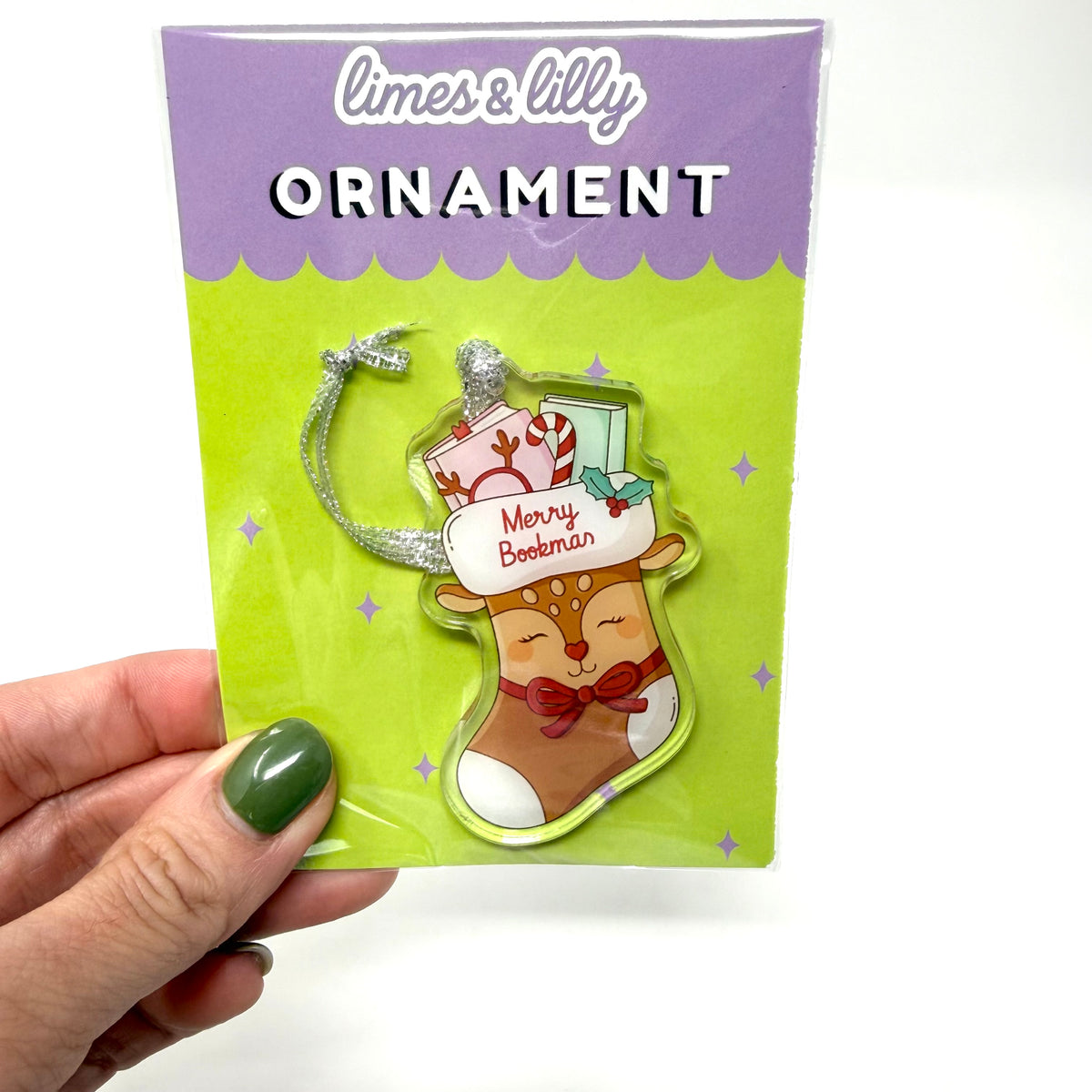 A hand with green nail polish holds a packaged Christmas ornament. The product is a stocking-shaped ornament featuring a sleeping reindeer with a red bow, a white hat reading 'Merry Botherman,' candy canes, and a silver crescent moon hanger.