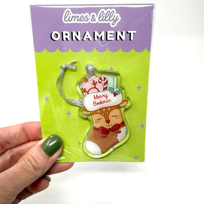 A hand with green nail polish holds a packaged Christmas ornament. The product is a stocking-shaped ornament featuring a sleeping reindeer with a red bow, a white hat reading 'Merry Botherman,' candy canes, and a silver crescent moon hanger.