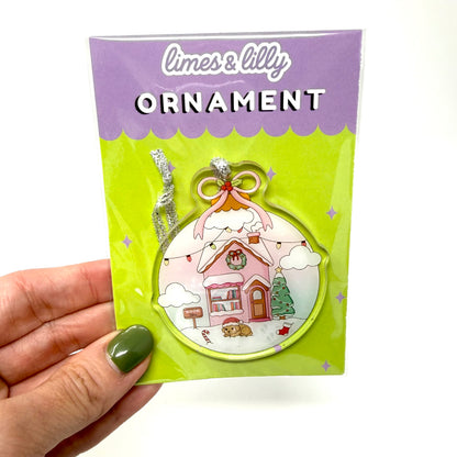A hand with olive green nail polish holds a packaged Christmas ornament. The product is a limes & lilly ornament featuring a whimsical pink house with a snowy roof, set against a bright lime green backing card with a purple header.