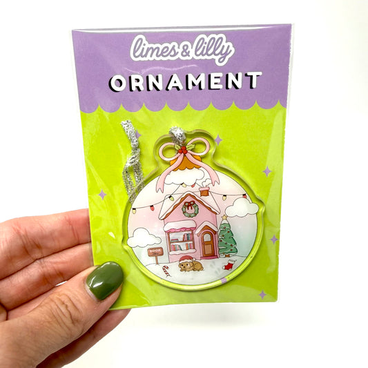 A hand with olive green nail polish holds a packaged Christmas ornament. The product is a limes & lilly ornament featuring a whimsical pink house with a snowy roof, set against a bright lime green backing card with a purple header.