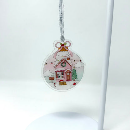 A round clear acrylic Christmas ornament featuring a whimsical pink house with a snowy roof, hanging from a silver string on a white background.