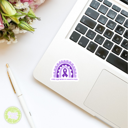 Laptop with a purple ribbon sticker on a white surface with flowers and a pen.