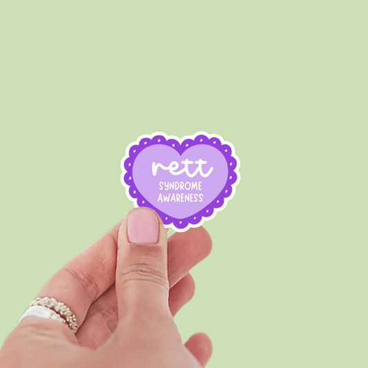 Hand holding a purple heart-shaped sticker with 'rett syndrome awareness' text on a light green background