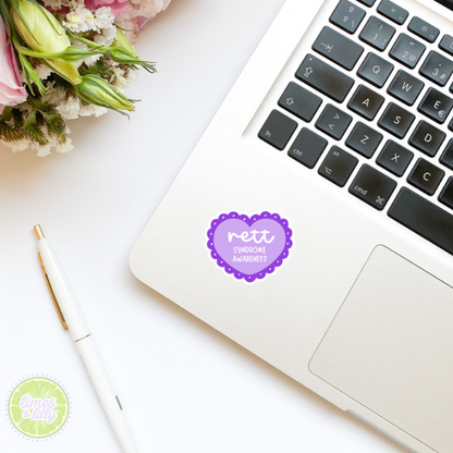 Laptop with a Rett Syndrome Awareness sticker on a white surface with flowers and a pen.
