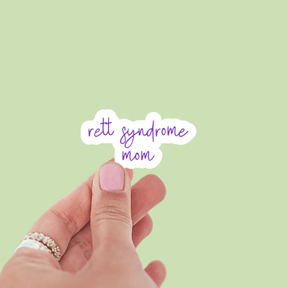 Hand holding a sticker with 'Rett Syndrome Mom' text on a light green background