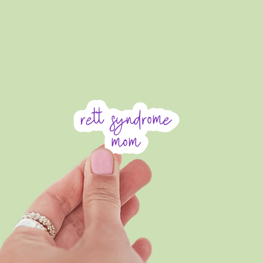 Hand holding a sticker with 'Rett Syndrome Mom' text on a light green background