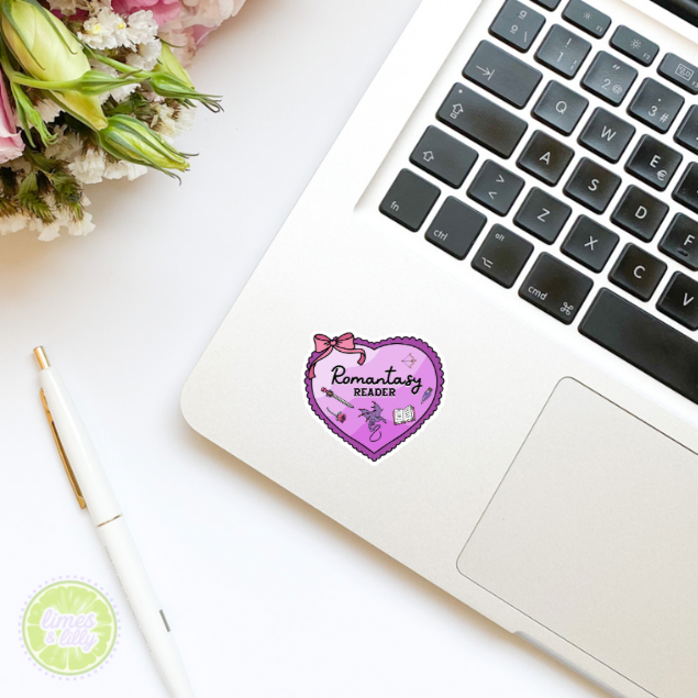 Laptop with a 'Romantic Reader' sticker on a white surface with flowers and a pen.