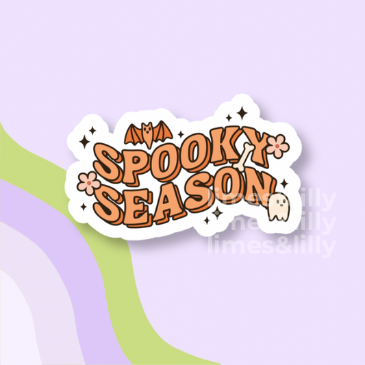A die-cut sticker with the words 'SPOOKY SEASON' in orange bubble letters, decorated with a bat, flowers, sparkles, and a small ghost on a white background, shown on a pastel purple backdrop with curved green and lavender stripes.