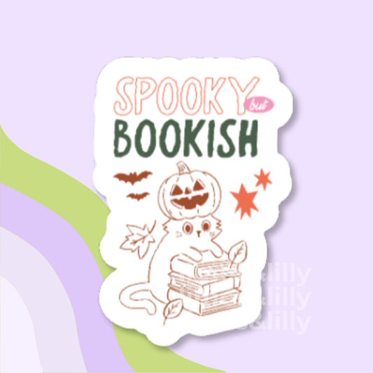 A die-cut sticker with a white background featuring a cute illustration of a cat reading books with a jack-o'-lantern head, plus the text 'SPOOKY but BOOKISH' in peach and dark green lettering. The sticker is centered on a pastel lavender background with abstract green and purple curved shapes.