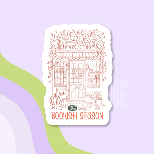 A die-cut sticker featuring a hand-drawn illustration of a book store storefront with pumpkins and autumn leaves, with the text 'The Bookish Season' on a light purple background with abstract green and lavender curved shapes.
