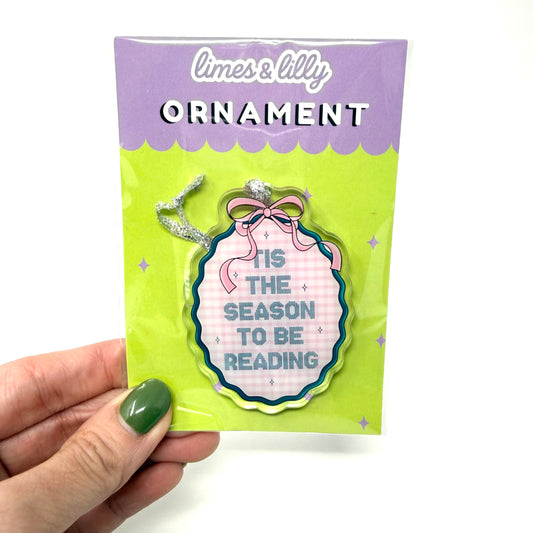 A hand with green nail polish holds a packaged acrylic ornament against a white background. The ornament is shaped like a book and has a pink gingham pattern with blue text reading 'IT'S THE SEASON TO BE READING'.