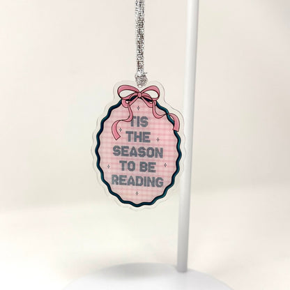 An acrylic hanging ornament with a scalloped edge, featuring a pink gingham pattern, a pink ribbon bow illustration, and the text 'IT'S THE SEASON TO BE READING' in navy blue. The ornament is suspended by a silver chain from a white display stand against a clean white background.