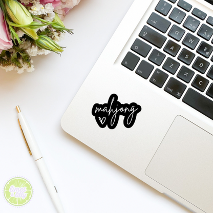 Laptop with 'mahjong' sticker on a white surface with flowers and a pen.